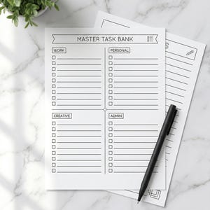 May include: White task management notepad with the text "MASTER TASK BANK" and sections for Work, Personal, Creative, and Admin tasks. A black pen rests on the notepad. The notepad is on a marble surface.