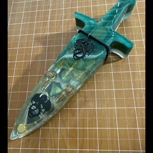 Puede incluir: Una daga decorativa encapsulada en resina transparente con detalles verdes y dorados. El mango y la hoja presentan objetos incrustados, incluyendo una calavera, dados y elementos decorativos. Un cord&oacute;n negro asegura la daga.