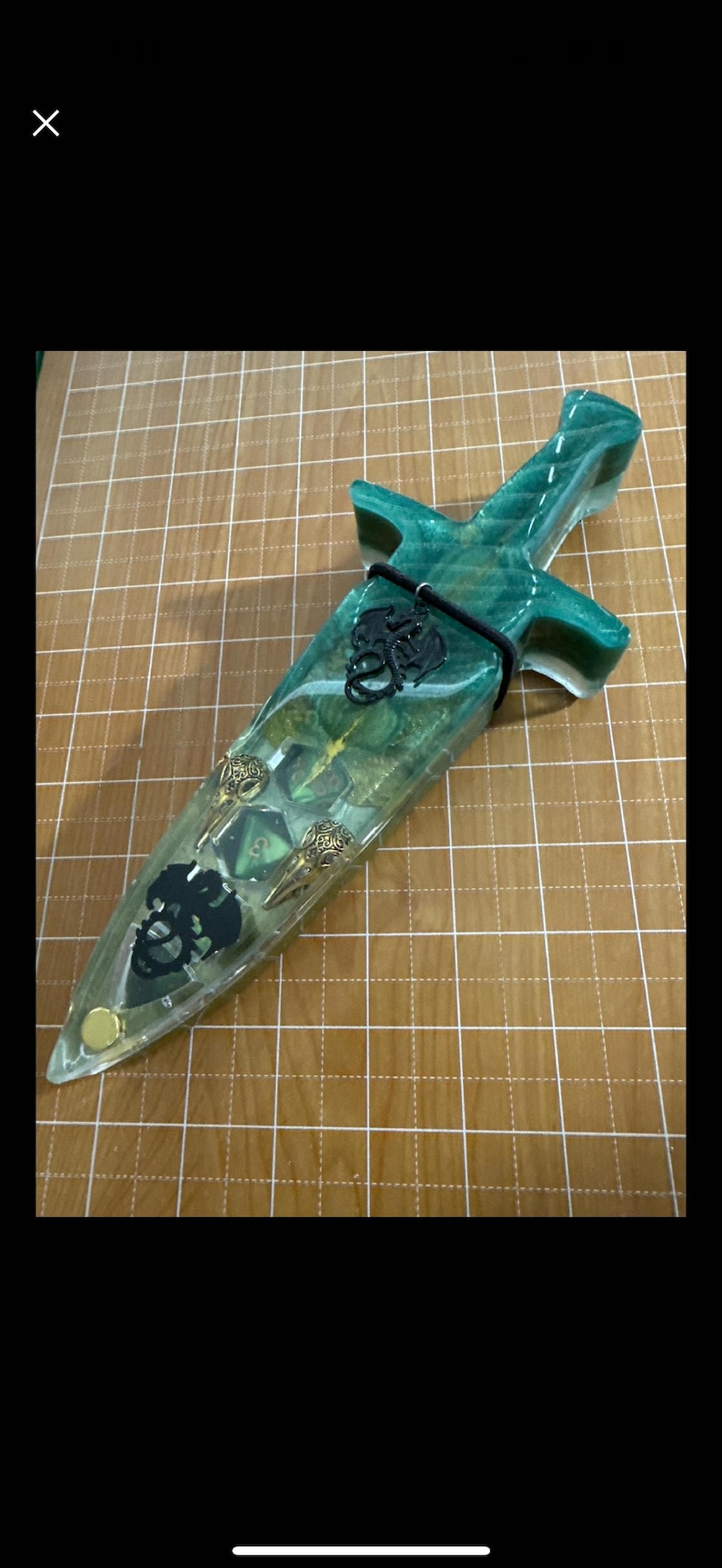 Puede incluir: Una daga decorativa encapsulada en resina transparente con detalles verdes y dorados. El mango y la hoja presentan objetos incrustados, incluyendo una calavera, dados y elementos decorativos. Un cord&oacute;n negro asegura la daga.