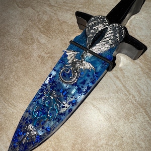 Puede incluir: Una daga decorativa con un mango negro y una hoja azul y transparente. La hoja est&aacute; adornada con adornos plateados en forma de drag&oacute;n y alas. La hoja est&aacute; llena de purpurina azul.