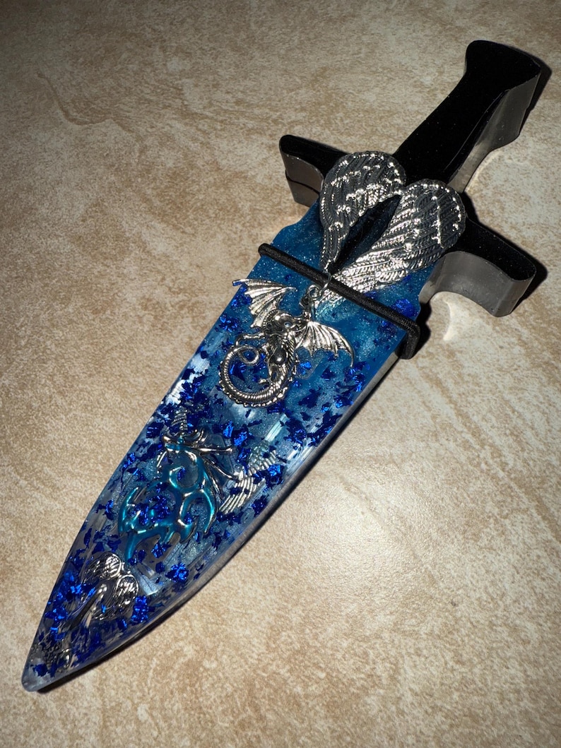 Puede incluir: Una daga decorativa con un mango negro y una hoja azul y transparente. La hoja est&aacute; adornada con adornos plateados en forma de drag&oacute;n y alas. La hoja est&aacute; llena de purpurina azul.