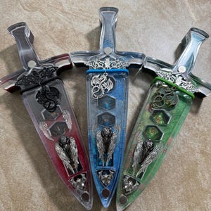 Puede incluir: Tres recipientes decorativos en forma de daga con mangos transparentes e interiores coloridos. Los recipientes tienen adornos plateados, incluyendo dise&ntilde;os de dragones y alas de &aacute;ngel. Los interiores son rojos, azules y verdes, cada uno con dados.