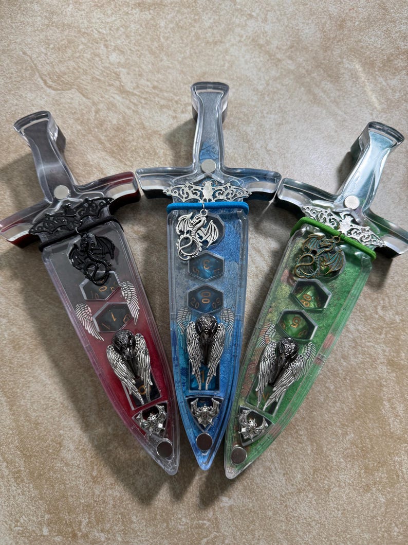 Puede incluir: Tres recipientes decorativos en forma de daga con mangos transparentes e interiores coloridos. Los recipientes tienen adornos plateados, incluyendo dise&ntilde;os de dragones y alas de &aacute;ngel. Los interiores son rojos, azules y verdes, cada uno con dados.