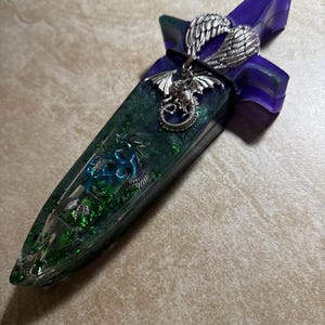 Puede incluir: Una daga decorativa con un mango morado y una hoja verde. La hoja est&aacute; incrustada con purpurina verde y un dise&ntilde;o de drag&oacute;n plateado. Un colgante de drag&oacute;n plateado y alas plateadas est&aacute;n unidos al mango.