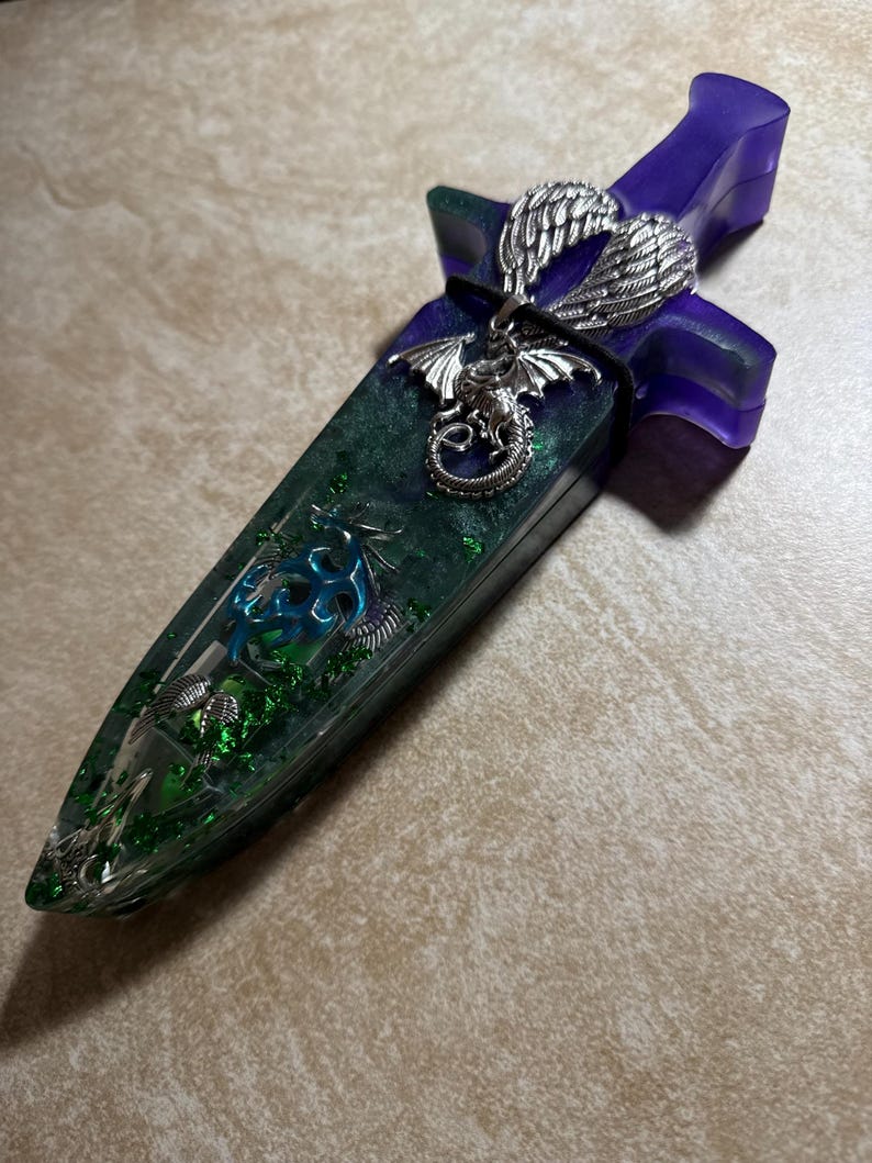 Puede incluir: Una daga decorativa con un mango morado y una hoja verde. La hoja est&aacute; incrustada con purpurina verde y un dise&ntilde;o de drag&oacute;n plateado. Un colgante de drag&oacute;n plateado y alas plateadas est&aacute;n unidos al mango.