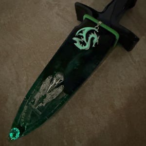 Puede incluir: Una daga decorativa con un mango negro y una vaina verde y negra. La vaina presenta un colgante de drag&oacute;n plateado, un dise&ntilde;o de calavera de p&aacute;jaro plateado y una gema verde brillante en la punta. La daga tiene un acento verde.