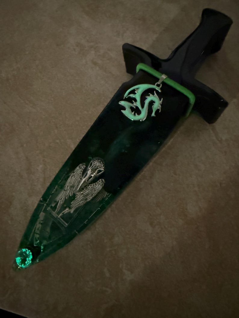 Puede incluir: Una daga decorativa con un mango negro y una vaina verde y negra. La vaina presenta un colgante de drag&oacute;n plateado, un dise&ntilde;o de calavera de p&aacute;jaro plateado y una gema verde brillante en la punta. La daga tiene un acento verde.