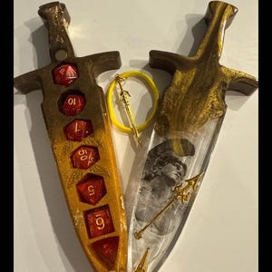 Puede incluir: Dos piezas decorativas de resina con forma de daga. Una es dorada y &aacute;mbar con dados rojos, la otra es transparente con un dise&ntilde;o dorado y una imagen en blanco y negro. Un anillo amarillo con un dije de espada dorada est&aacute; entre ellas.
