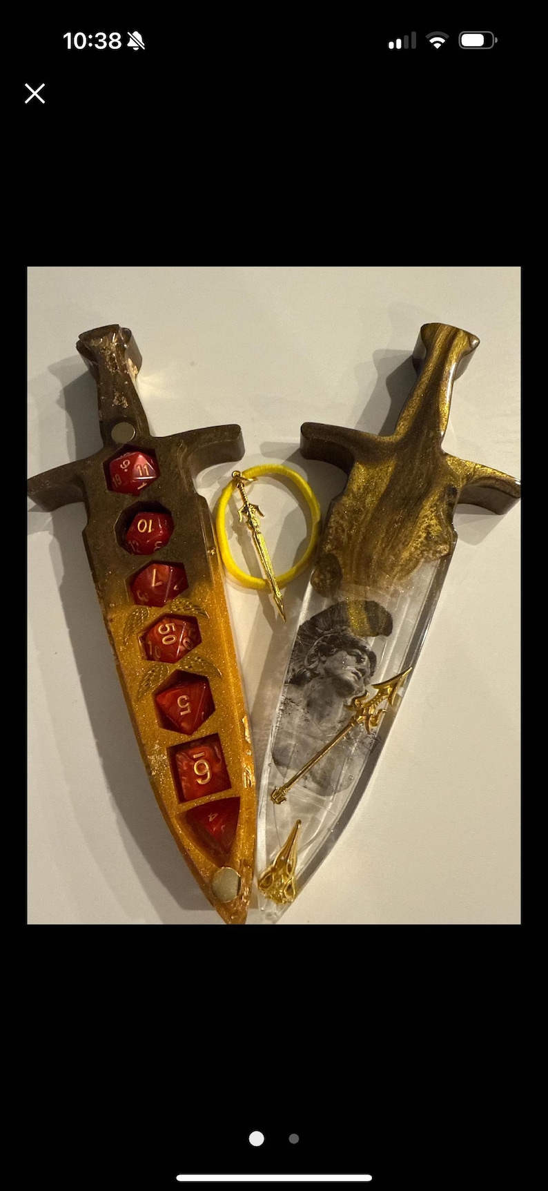 Puede incluir: Dos piezas decorativas de resina con forma de daga. Una es dorada y &aacute;mbar con dados rojos, la otra es transparente con un dise&ntilde;o dorado y una imagen en blanco y negro. Un anillo amarillo con un dije de espada dorada est&aacute; entre ellas.