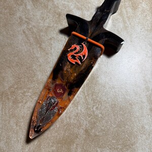 Puede incluir: Una daga decorativa con mango negro y hoja encapsulada en resina transparente. La hoja presenta un colgante de drag&oacute;n rojo, escamas met&aacute;licas incrustadas y un emblema rojo. La daga mide aproximadamente 25 cm de largo.