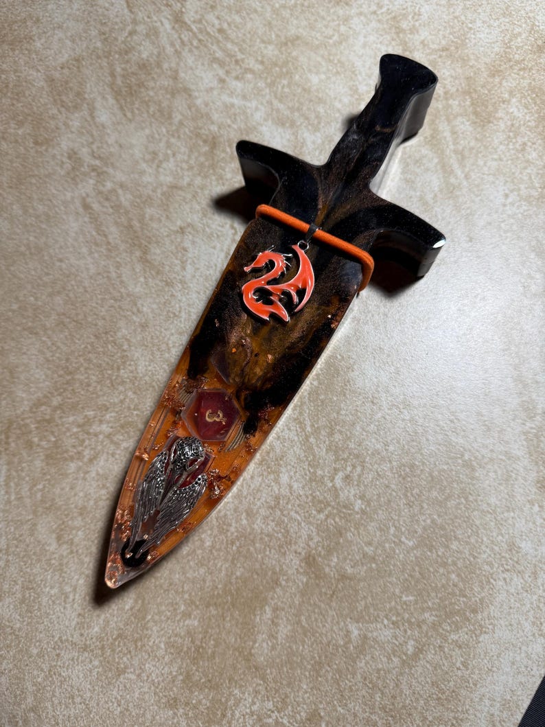 Puede incluir: Una daga decorativa con mango negro y hoja encapsulada en resina transparente. La hoja presenta un colgante de drag&oacute;n rojo, escamas met&aacute;licas incrustadas y un emblema rojo. La daga mide aproximadamente 25 cm de largo.