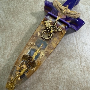 Puede incluir: Una daga decorativa con un mango morado y una hoja transparente llena de copos de oro y dados. La daga presenta adornos dorados de drag&oacute;n y &aacute;guila, a&ntilde;adiendo un tema de fantas&iacute;a.