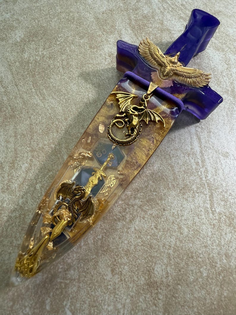 Puede incluir: Una daga decorativa con un mango morado y una hoja transparente llena de copos de oro y dados. La daga presenta adornos dorados de drag&oacute;n y &aacute;guila, a&ntilde;adiendo un tema de fantas&iacute;a.
