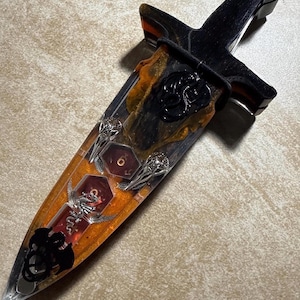 Puede incluir: Una daga decorativa con mango negro y hoja encapsulada en resina transparente con detalles en naranja y negro. La hoja presenta detalles de dados, calaveras y dragones. La daga mide aproximadamente 25 cm de largo.