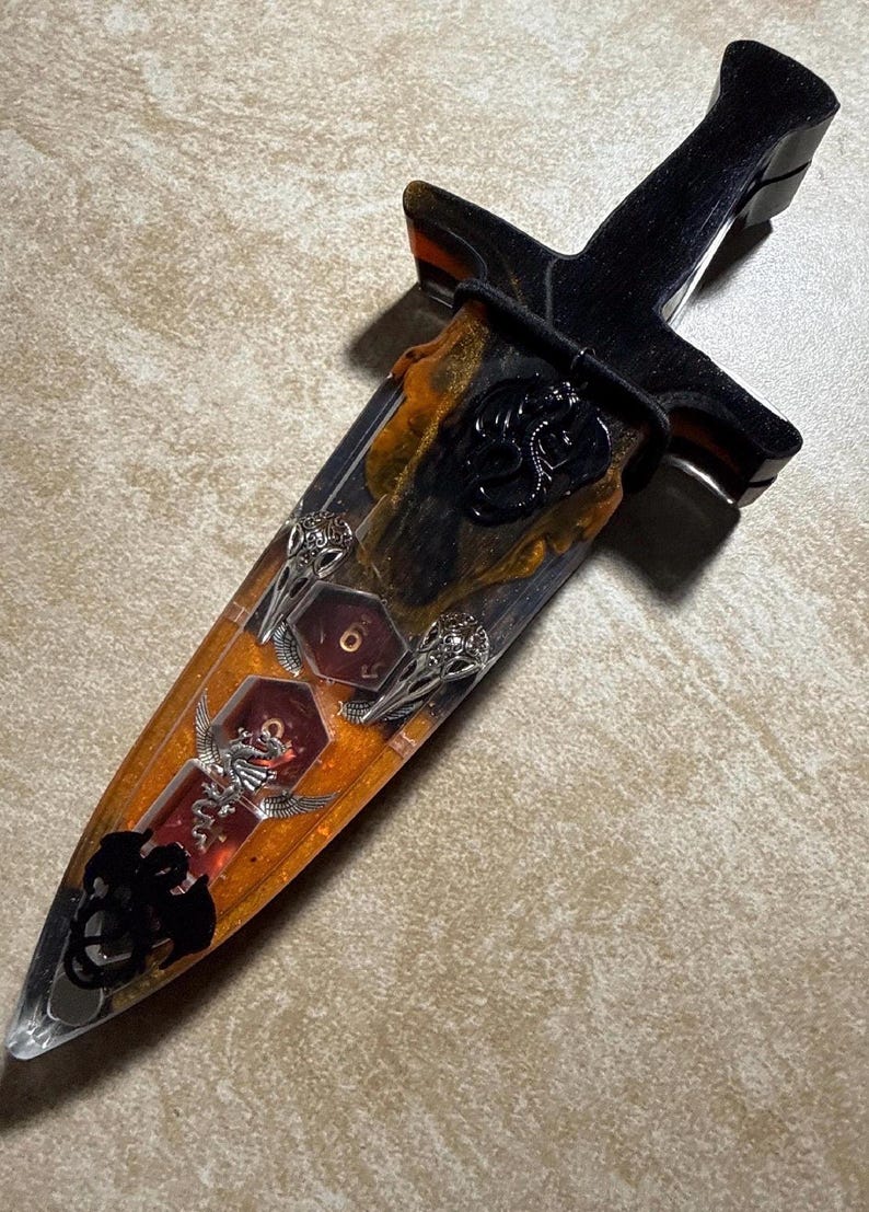 Puede incluir: Una daga decorativa con mango negro y hoja encapsulada en resina transparente con detalles en naranja y negro. La hoja presenta detalles de dados, calaveras y dragones. La daga mide aproximadamente 25 cm de largo.