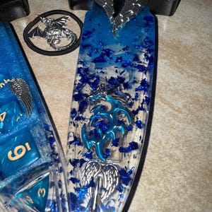 Puede incluir: Un conjunto de tem&aacute;tica fant&aacute;stica con una espada de resina azul y un juego de dados. La espada tiene un mango transparente con detalles azules y un dise&ntilde;o de alas plateadas. El juego de dados incluye dados azules con n&uacute;meros dorados y una goma para el pelo de drag&oacute;n.
