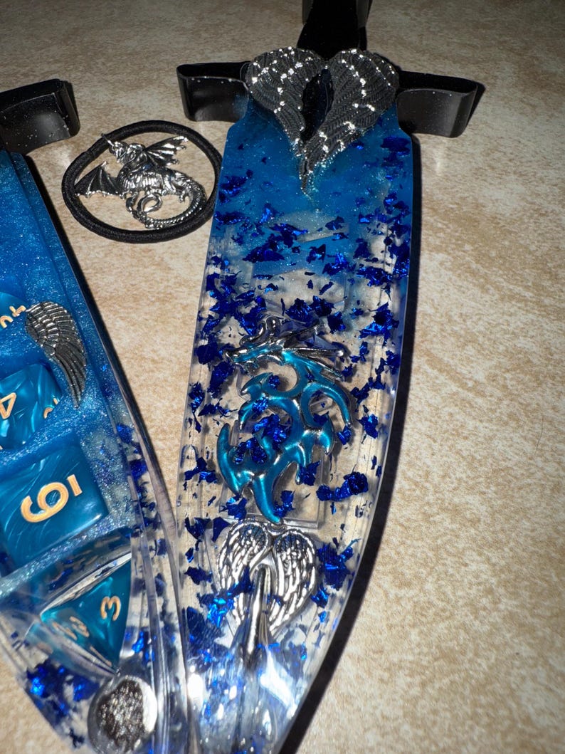 Puede incluir: Un conjunto de tem&aacute;tica fant&aacute;stica con una espada de resina azul y un juego de dados. La espada tiene un mango transparente con detalles azules y un dise&ntilde;o de alas plateadas. El juego de dados incluye dados azules con n&uacute;meros dorados y una goma para el pelo de drag&oacute;n.