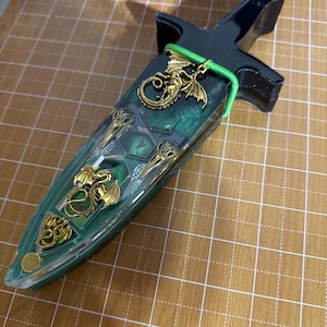 Puede incluir: Una caja decorativa en forma de daga con un mango negro y una hoja verde y transparente. La hoja est&aacute; adornada con adornos de dragones dorados y un dije de drag&oacute;n dorado est&aacute; sujeto al mango con una banda verde. La caja est&aacute; sobre una superficie de cuadr&iacute;cula marr&oacute;n.