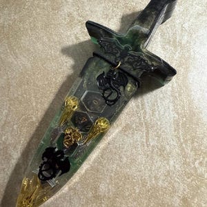 Puede incluir: Una daga decorativa con mango y hoja verdes y negros. La hoja est&aacute; llena de dados y detalles dorados. Un amuleto de drag&oacute;n negro cuelga de la empu&ntilde;adura. La daga tiene un tema de fantas&iacute;a.