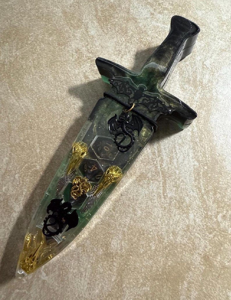 Puede incluir: Una daga decorativa con mango y hoja verdes y negros. La hoja est&aacute; llena de dados y detalles dorados. Un amuleto de drag&oacute;n negro cuelga de la empu&ntilde;adura. La daga tiene un tema de fantas&iacute;a.