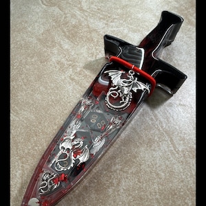Puede incluir: Una daga decorativa con una hoja de resina transparente llena de purpurina roja y dados. El mango es negro y rojo, con un dije de drag&oacute;n plateado y detalles rojos. La daga mide aproximadamente 25 cm de largo.