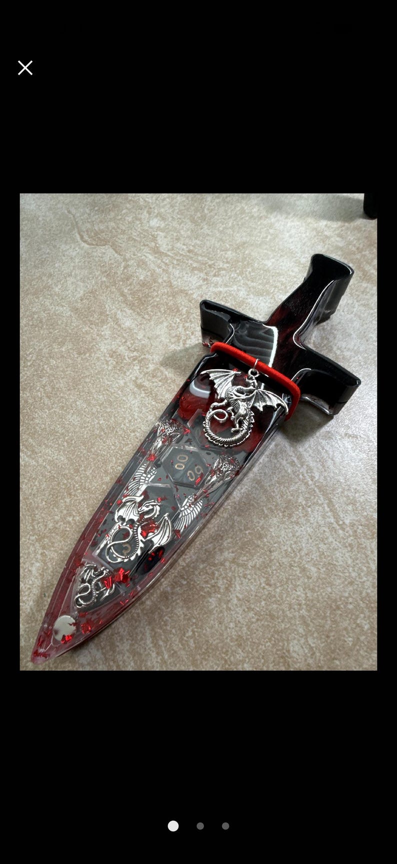 Puede incluir: Una daga decorativa con una hoja de resina transparente llena de purpurina roja y dados. El mango es negro y rojo, con un dije de drag&oacute;n plateado y detalles rojos. La daga mide aproximadamente 25 cm de largo.