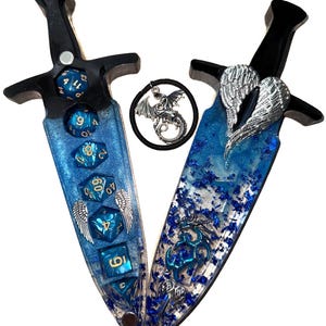 May include: Two decorative dagger-shaped objects with black handles and blue resin blades. The blades are embedded with blue dice and silver wing accents. A circular charm with a dragon is between the daggers.