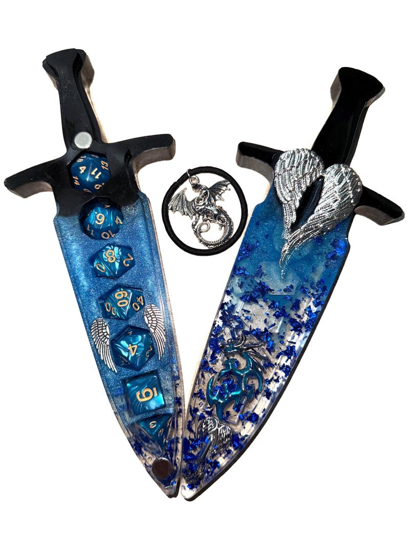 Puede incluir: Dos objetos decorativos en forma de daga con mangos negros y hojas de resina azul. Las hojas est&aacute;n incrustadas con dados azules y detalles de alas plateadas. Un amuleto circular con un drag&oacute;n est&aacute; entre las dagas.