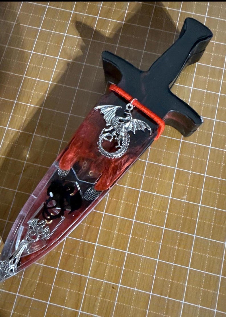 Puede incluir: Una daga decorativa con un mango negro y una hoja transparente llena de resina roja y negra. Un dije de drag&oacute;n plateado cuelga del mango. La hoja presenta adornos plateados adicionales.