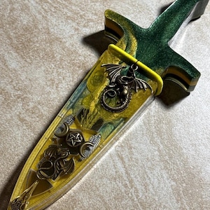 Puede incluir: Una daga decorativa con un mango verde y amarillo y una vaina a juego. La vaina est&aacute; adornada con un amuleto de drag&oacute;n de bronce y otros s&iacute;mbolos m&iacute;sticos. La daga mide aproximadamente 25 cm de largo.
