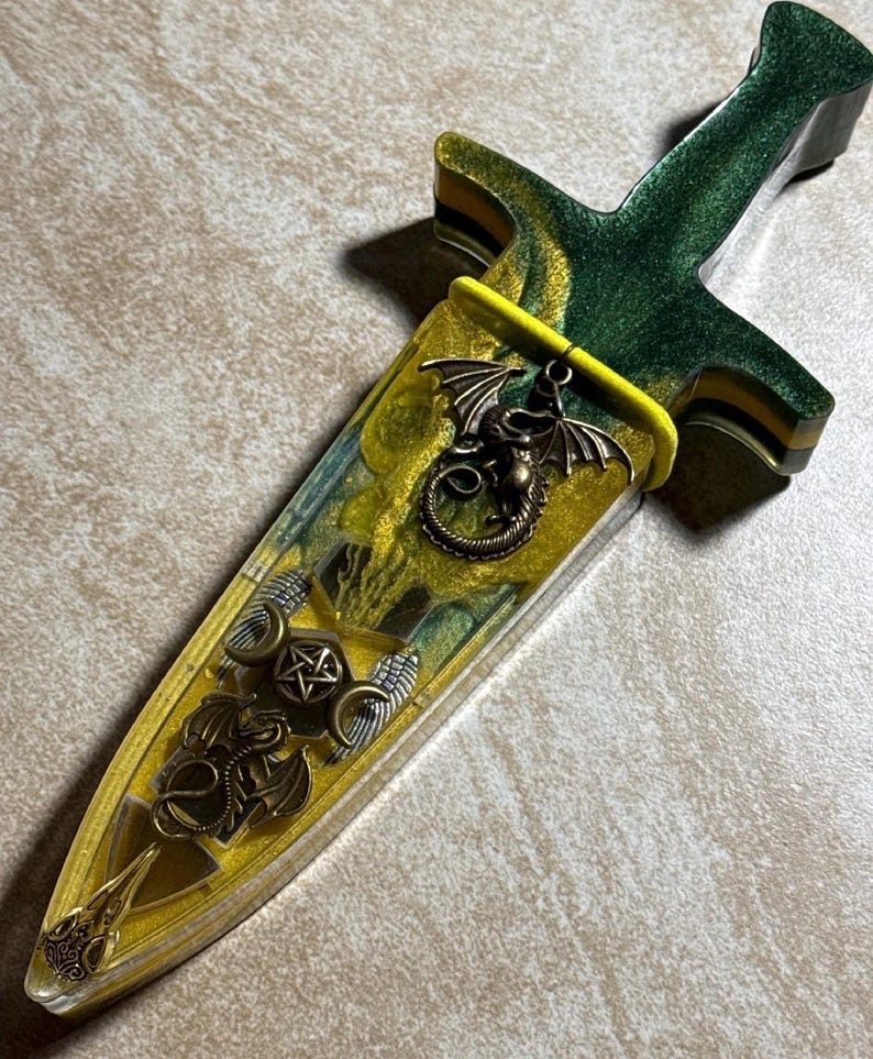 Puede incluir: Una daga decorativa con un mango verde y amarillo y una vaina a juego. La vaina est&aacute; adornada con un amuleto de drag&oacute;n de bronce y otros s&iacute;mbolos m&iacute;sticos. La daga mide aproximadamente 25 cm de largo.