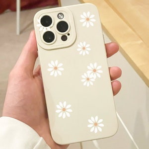 Peut inclure: Une coque de téléphone beige ornée de motifs de fleurs de marguerite blanches. L'étui a une découpe pour l'appareil photo et est tenu dans une main. Les marguerites sont espacées uniformément sur la surface.