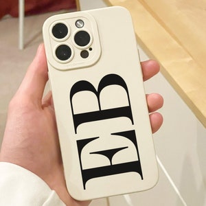 May include: A cream-colored phone case with a black monogram of the letters "RB". The case has a camera cutout with four lenses and a flash. The phone case is held in a person's hand.