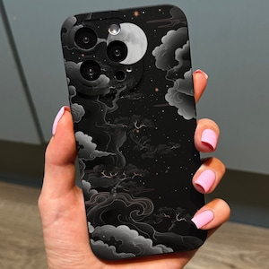 Puede incluir: Funda de teléfono negra con un diseño de cielo nocturno con nubes y una luna. La funda tiene un acabado brillante y se sostiene en una mano con esmalte de uñas rosa. El diseño incluye estrellas y una luna creciente.