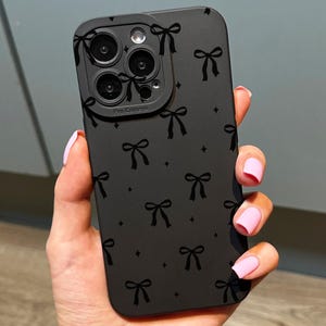 May include: Black phone case with a bow pattern. The case has a matte finish and features a repeating design of small bows and tiny star shapes. The camera lens area is visible.