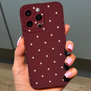 May include: A burgundy phone case with a white polka dot pattern. The case has a camera lens protector. The background is a neutral grey and wood-tone surface.