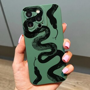 以下が含まれることがあります： セージグリーンのスマートフォンケース。背面には2匹のヘビが絡み合う黒いグラフィックイラストが描かれています。鱗の模様や舌まで詳細に表現されており、カメラレンズとフラッシュモジュール用の切り抜き穴が配置されています。