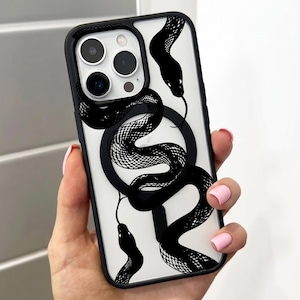 Può includere: Una custodia trasparente per smartphone con bordo nero e una stampa grafica nera di un serpente che si avvolge sul retro. Il design presenta un anello magnetico circolare al centro e un ritaglio per la fotocamera a tripla lente in alto.