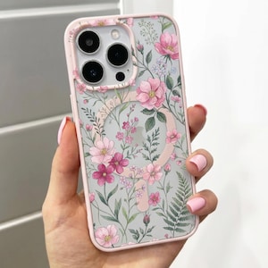 Pode incluir: Uma capa de telemóvel transparente com rebordo rosa, apresentando um padrão floral delicado de flores cor-de-rosa, folhas verdes e fetos. A capa inclui um design de anel magnético circular na parte traseira, rodeando o logótipo Apple.