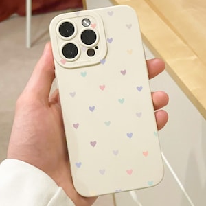 May include: A cream phone case decorated with a pattern of small, pastel-coloured hearts. The case has a protective camera lens surround. The case is held in a hand, showing the design and fit.