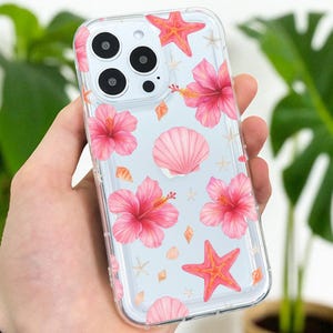 Op de afbeelding: Transparante telefoonhoesje met een tropisch ontwerp met roze hibiscusbloemen, zeesterren en schelpen op een lichtblauwe achtergrond. De hoes wordt in een hand gehouden.