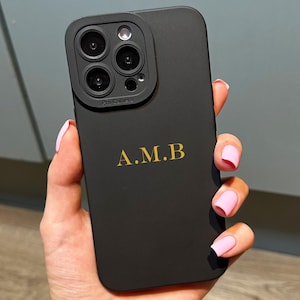 May include: Black phone case with a gold monogram of the letters A.M.B. The case has a matte finish and a camera lens protector. The phone case is held in a hand with pink nail polish.