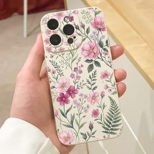May include: A floral phone case featuring a cream background with pink and purple flowers, green leaves, and a fern. The case has cutouts for the phone's camera and buttons.