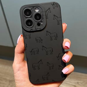 May include: Black phone case featuring a repeating pattern of donkey illustrations. The case has a camera lens protector. The phone case is held in a hand with pink nail polish. The phone case is a solid black colour.
