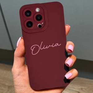 May include: A burgundy phone case with a camera cutout and the name "Olivia" in a cursive font. The case is held in a hand with pink nail polish. The phone case is designed to protect the phone.