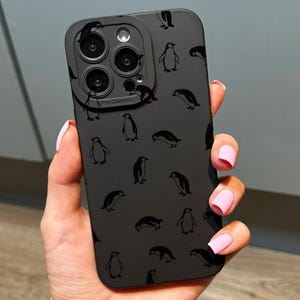 May include: Black phone case with a repeating pattern of penguin designs. The case includes a camera lens protector. The phone case is held in a hand with pink nail polish. The phone case is designed to fit a smartphone.