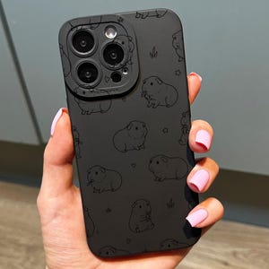 May include: Black phone case with a repeating pattern of guinea pig illustrations. The case has a matte finish and a camera lens protector. The design includes small stars and blades of grass. The phone case is held in a hand.