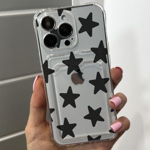 May include: Clear phone case with black star pattern. The case has a built-in card holder and is designed to fit a smartphone. The case is held in a hand with pink nail polish.
