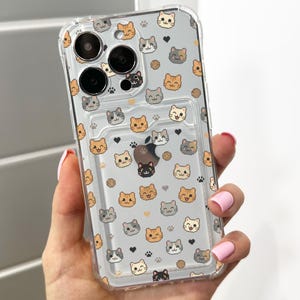Puede incluir: Funda transparente para teléfono con un patrón repetido de caras de gatos de dibujos animados en varios colores. La funda tiene un tarjetero incorporado y se sostiene en una mano con esmalte de uñas rosa.