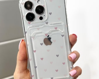 May include: Clear phone case with a heart pattern in shades of pink and gray. The case has a cardholder on the back and a cutout for the Apple logo. The phone case is designed to fit a smartphone.